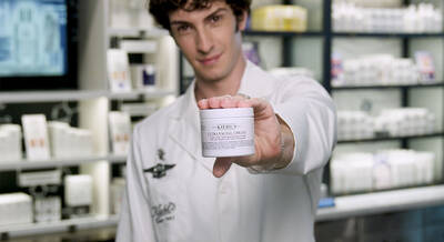 A person in a lab coat holds up a jar of Kiehl's Ultra Facial Cream, standing in front of a display of skincare products.