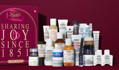 Boite cadeau du Calendrier de l'Avent Kiehl's en rouge profond avec texte dore, exposee a cote de 24 mini-produits de soin, dont la Creme Ultra Faciale, le Tonique au Calendula, le Serum au Retinol et le Baume a Levres n°1. Un graphique en spray rose sur le cote met en evidence la valeur de l'ensemble.