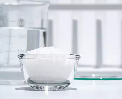 A small glass bowl filled with white granules sits on a table next to a measuring beaker containing clear liquid. In the background, a white surface provides a simple backdrop.