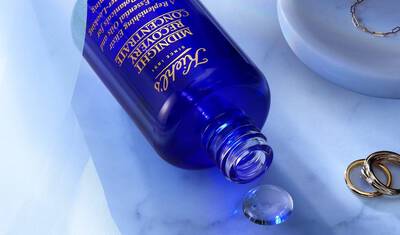 A blue bottle of Tatcha Midnight Concentrate is lying on a marble surface, with a small drop of the product spilling out. Nearby, there are a few gold rings and a white circular tray.