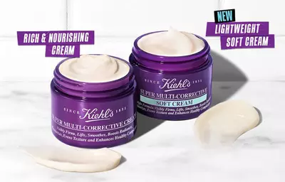 Two jars of Kiehl's Super Multi-Corrective Cream are displayed on a marble surface. One jar is labeled "Rich & Nourishing Cream" and the other "New Lightweight Soft Cream," with cream texture visible inside the jars.