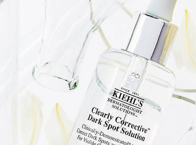 Flacon de solution Correctrice pour taches sombres de Kiehl's, entoure de fleurs blanches et de verre, sur fond clair.