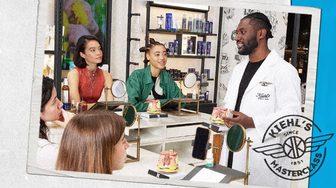 Een man in een Kiehl's labjas leidt een masterclass met verschillende deelnemers aan een tafel met spiegels en modellen, terwijl hij iets uitlegt, tegen een achtergrond van productschappen en een Kiehl's Masterclass-logo.