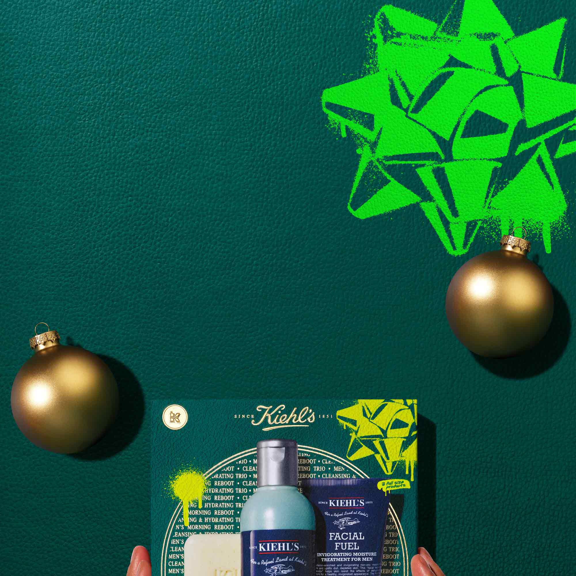 A graphic, set against a dark green textured background, features three Kiehl’s products and a gift box. The top displays a bar of "KIEHL’S 'ULTIMATE MAN' BODY SCRUB SOAP," a blue bottle of "KIEHL’S FACIAL FUEL ENERGIZING FACE WASH," and a dark blue tube of "KIEHL’S FACIAL FUEL MOISTURIZER." Two gold spherical ornaments are also present. Below these, a dark teal Kiehl’s gift box showcases the same three products, decorated with bright green graphic paint splatters and a bow design.