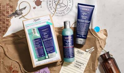 A flat lay image featuring Kiehl's Facial Fuel skincare products, including a bottle of Energizing Toning Wash and a tube of Moisturizer Treatment. The products are displayed on a textured, brown paper background, alongside a vintage-style label and blue sticky note.