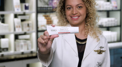 A person with curly hair holds a tube of Kiehl's Barrier Cream, smiling in a store filled with skincare products.