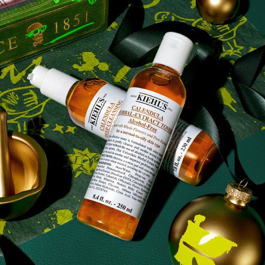 Kiehl's Calendula Herbal-Extract Toner en serum gerangschikt met gouden kerstornamenten en groen lint op een feestelijke donkergroene achtergrond.