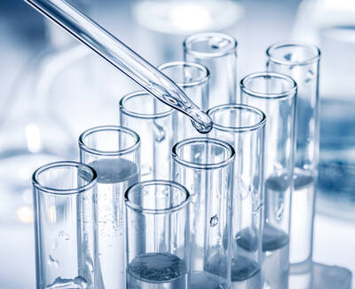 A pipette is dropping a liquid into a row of clear glass test tubes filled with water or a similar substance, set against a blurred laboratory background.