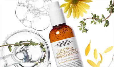 Alt text: A bottle of Kiehl's Calendula Deep Cleansing Foaming Face Wash surrounded by bubbles, flower petals, and herbs on a white background.