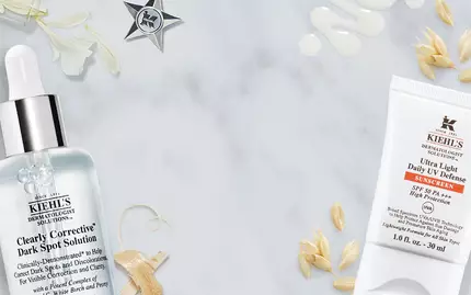 Alt-text in Dutch: "Twee Kiehl's huidverzorgingsproducten op een marmeren ondergrond, inclusief een serum en een creme, omgeven door natuurlijke ingredienten zoals haver."