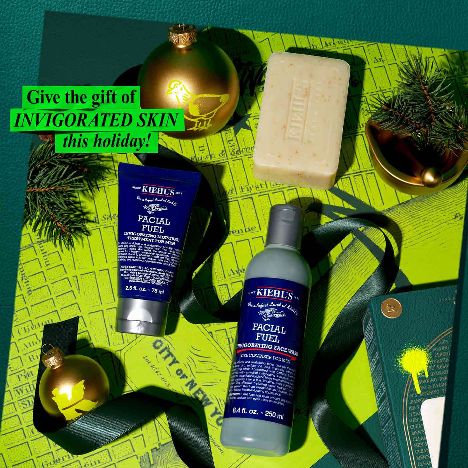 Een feestelijke afbeelding tegen een groene achtergrond met een landkaartpatroon toont drie Kiehl's-producten: een donkerblauwe tube "KIEHL'S FACIAL FUEL MOISTURIZER", een blauwe fles "KIEHL'S FACIAL FUEL ENERGIZING FACE WASH" en een reep "KIEHL'S 'ULTIMATE MAN' BODY SCRUB SOAP". Twee gouden kerstornamenten, een met een geel vogelmotief en een ander met een geel "K"-logo, maken deel uit van de opstelling, samen met donker lint en dennentakken. Rechtsonder is een gedeeltelijk beeld van een groene Kiehl's-cadeaudoos zichtbaar. Felgroene tekst linksboven luidt "Geef deze feestdagen de INVIGORATED SKIN cadeau!