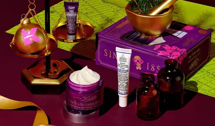 Un graphique festif, sur un fond rouge fonce avec un motif de carte verte, presente plusieurs produits Kiehl's et des decorations de vacances. Une balance doree est mise en evidence, avec un ornement spherique dore comportant un logo "K" rose d'un cote et un petit tube violet de "Kiehl's Super Multi-Corrective Eye Zone Treatment" de l'autre. Au premier plan, un pot violet ouvert de "Kiehl's Super Multi-Corrective Cream" avec de la creme visible est presente. Un tube blanc et violet de "Kiehl's Retinol Skin-Renewing Daily Micro-Dose Serum" se trouve a proximite. A l'arriere-plan, un mortier et un pilon dores avec des branches de pin et un coffret cadeau violet avec "SINCE 1851" et un motif de tete de mort sont visibles. Deux bouteilles en verre ambre et un ruban dore completent l'arrangement festif.