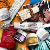 Een assortiment van Kiehl's huidverzorgingsproducten artistiek gerangschikt op een levendige oranje achtergrond met het Vrijheidsbeeld.
